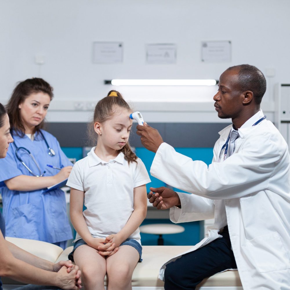 African american pediatrician doctor measuring temperature of young girl using medical infrared thermometer during appointment in hospital office. Therapist man explaining healthcare treatment