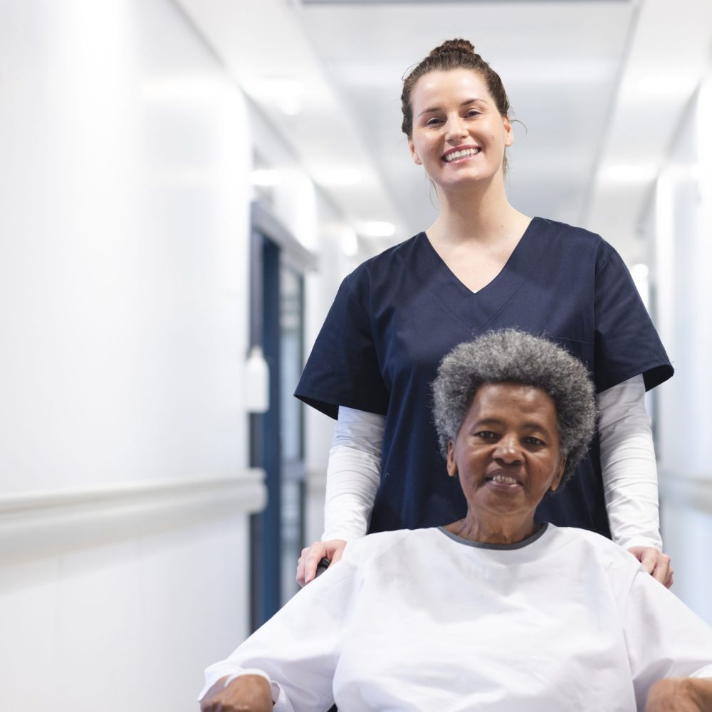 Happy diverse female doctor walking with senior female patient in wheelchair in hospital corridor. Medicine, healthcare and medical services, unaltered.