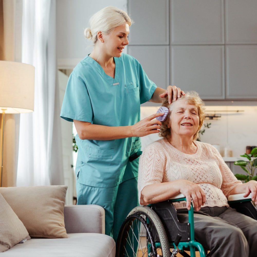 Nurse caring for a senior woman in a wheelchair at home, brushing her hair with compassion and warmth. The image shows the bond between caregiver and patient